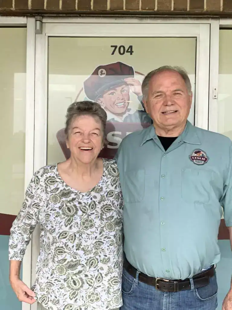 An older couple stands in front of a storefront with the number 704. The man wears a blue shirt, and the woman wears a floral blouse.