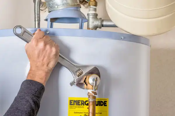 A person uses a wrench to tighten a pipe on a water heater. An "Energy Guide" label is visible on the heater.